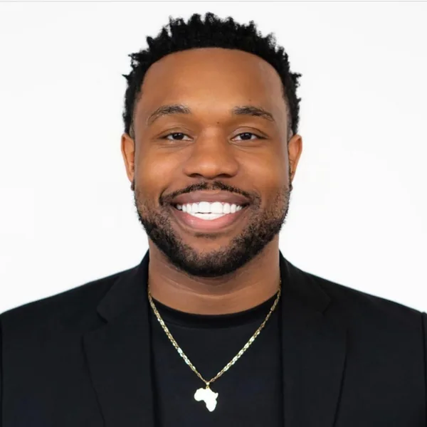 Professional headshot of Bakari Akil, Managing Director of Graves Hall Capital, smiling in a black blazer and black crew-neck shirt with a gold Africa pendant necklace against a white background
