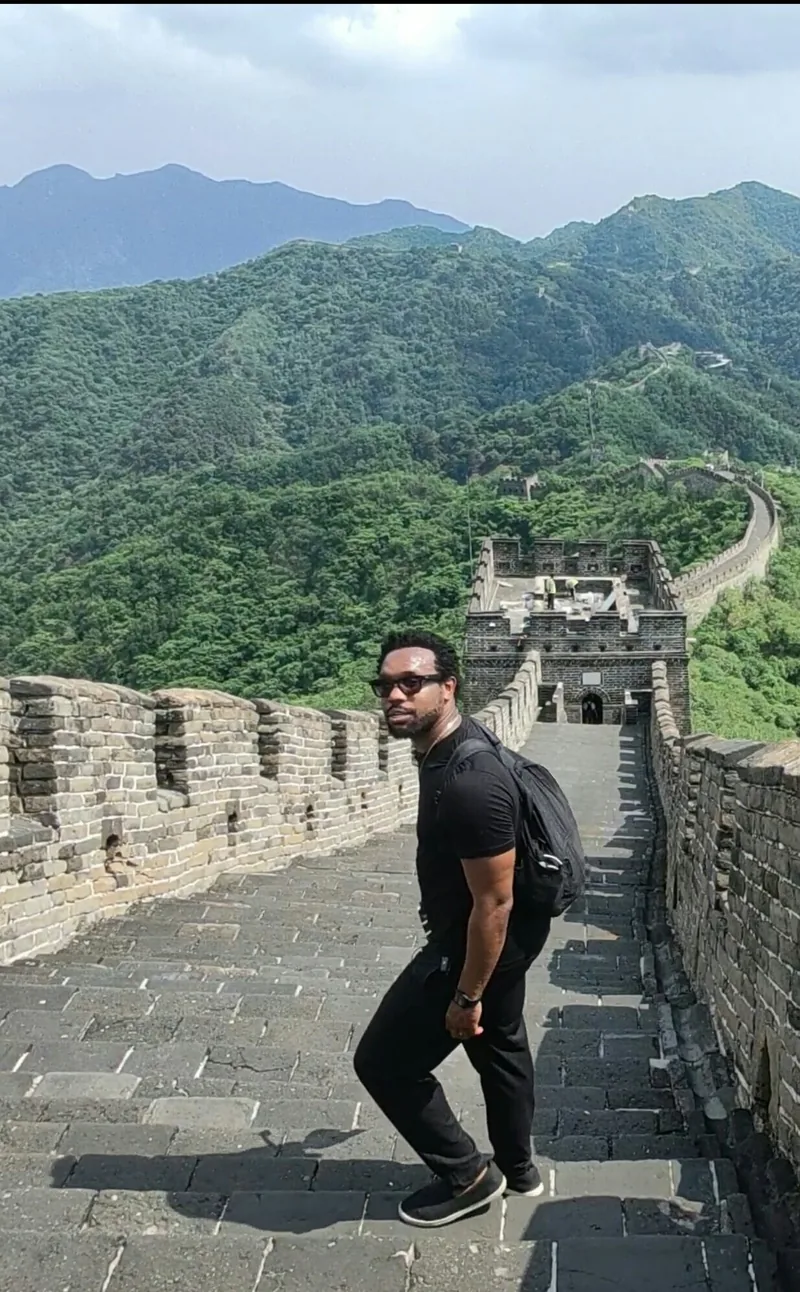 Bakari Akil climbing the Great Wall of China in sunglasses and a black polo, looking back at the camera with green mountains and watchtowers stretching into the distance behind him