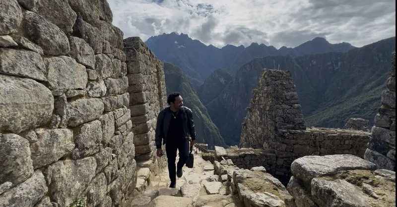 Bakari Akil walking through the ancient stone ruins of Machu Picchu, Peru, dressed in all black, the Andes mountains and misty cloud forest dropping away behind the Inca citadel