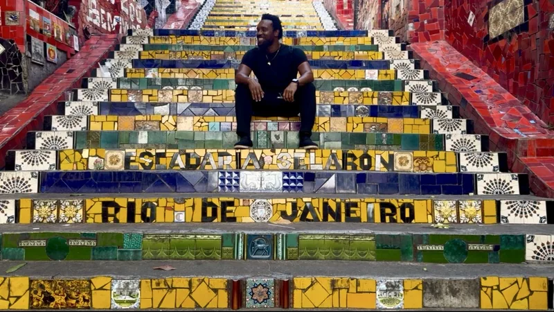 Bakari Akil standing on the colorful mosaic-tiled Escadaria Selarón steps in Rio de Janeiro, Brazil, tiles spelling out RIO DE JANEIRO in bright yellow, red, green, and blue around him