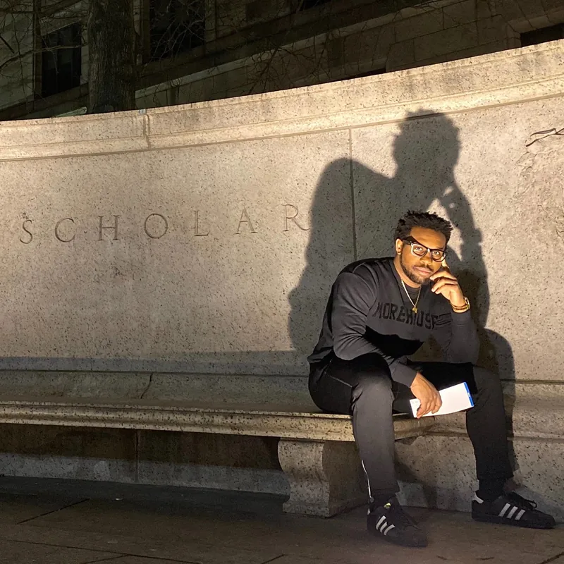Bakari Akil sitting on a stone bench beneath the word SCHOLAR carved into a university wall, wearing a Morehouse College sweatshirt and glasses, reading a book at night — author photo for his forthcoming memoir Paper Ceiling