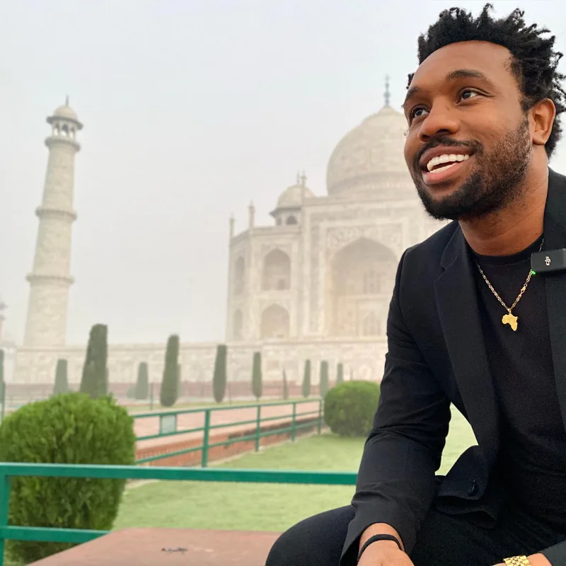 Bakari Akil smiling in a black blazer and gold Africa pendant necklace seated in front of the Taj Mahal in Agra, India, the white marble dome and minarets hazy in the morning light behind him