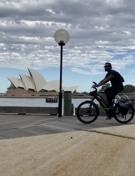 Bakari Akil riding a bicycle along the harbour boardwalk in Sydney, Australia, dressed in all black with sunglasses, the white sails of the Sydney Opera House across the water behind him