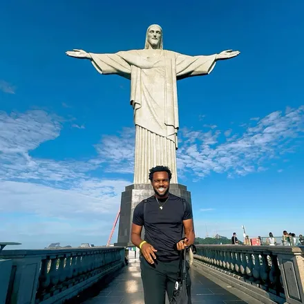 Bakari Akil smiling in a black t-shirt and gold Africa pendant necklace at the base of Christ the Redeemer on Corcovado mountain in Rio de Janeiro, Brazil, the statue's outstretched arms spanning the blue sky above him