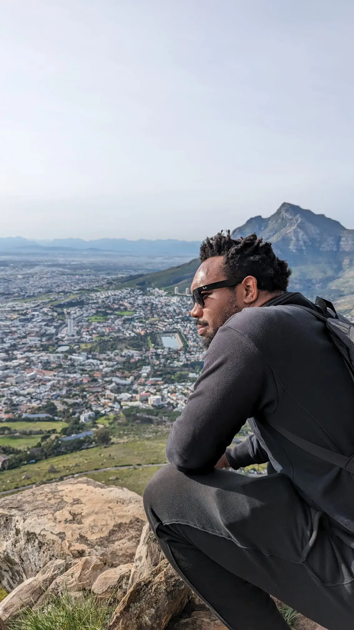 Bakari Akil seated on a rocky summit overlooking the city of Cape Town, South Africa, wearing sunglasses and a black sweater with a backpack, Table Mountain and the Atlantic coastline stretching out below