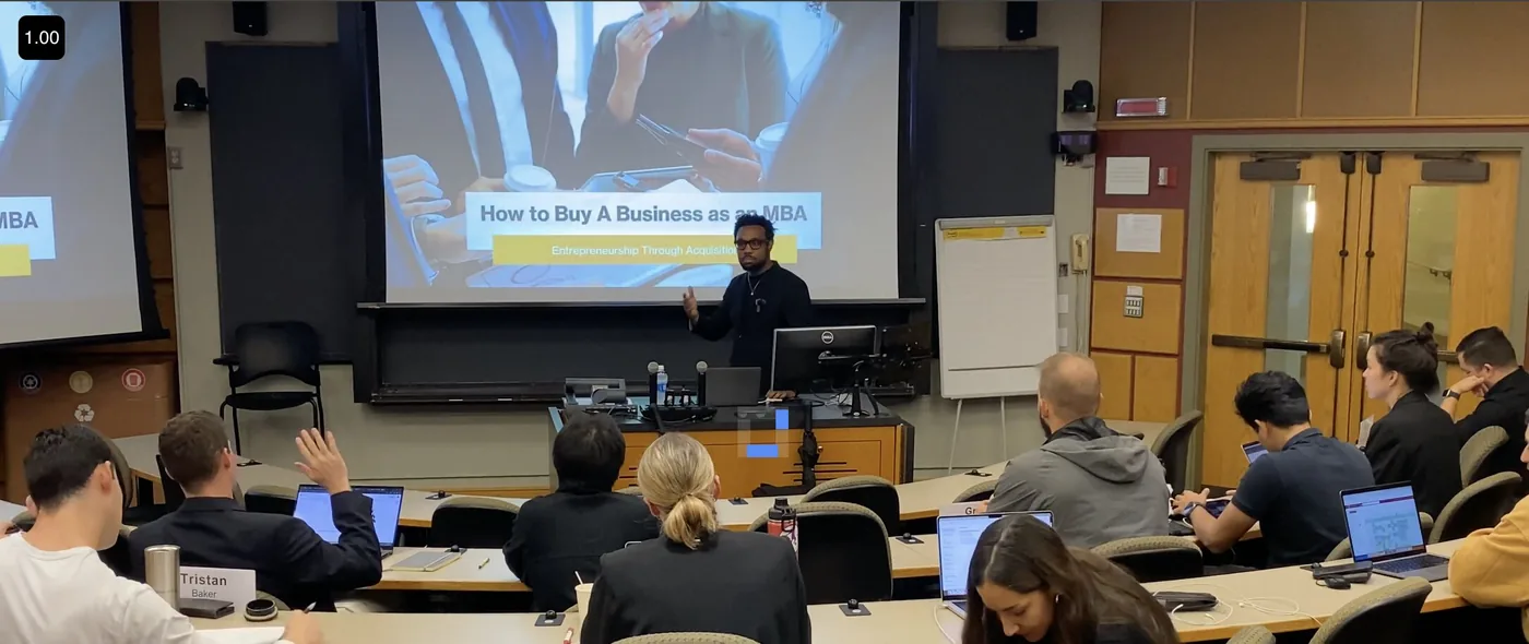Wide-angle view of Bakari Akil teaching How to Buy a Business to a full MBA classroom, gesturing at a projection screen while students take notes on laptops