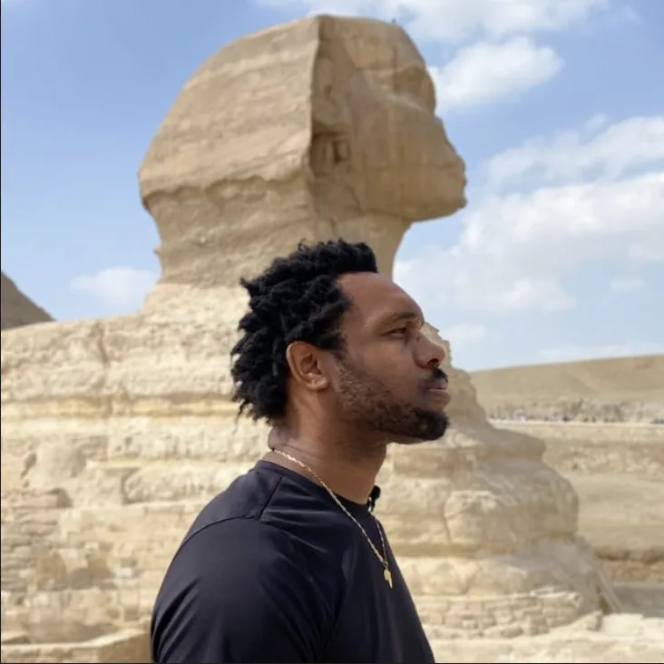 Bakari Akil in profile beside the Great Sphinx of Giza, Egypt — his silhouette mirroring the Sphinx's, wearing a black t-shirt and gold chain with the Giza pyramids and blue sky behind him