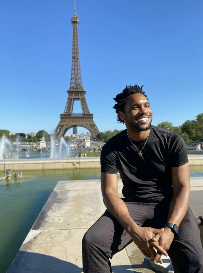 Bakari Akil seated at the Trocadéro fountains in Paris smiling in a black t-shirt and gold chain, the Eiffel Tower rising behind him against a clear blue sky