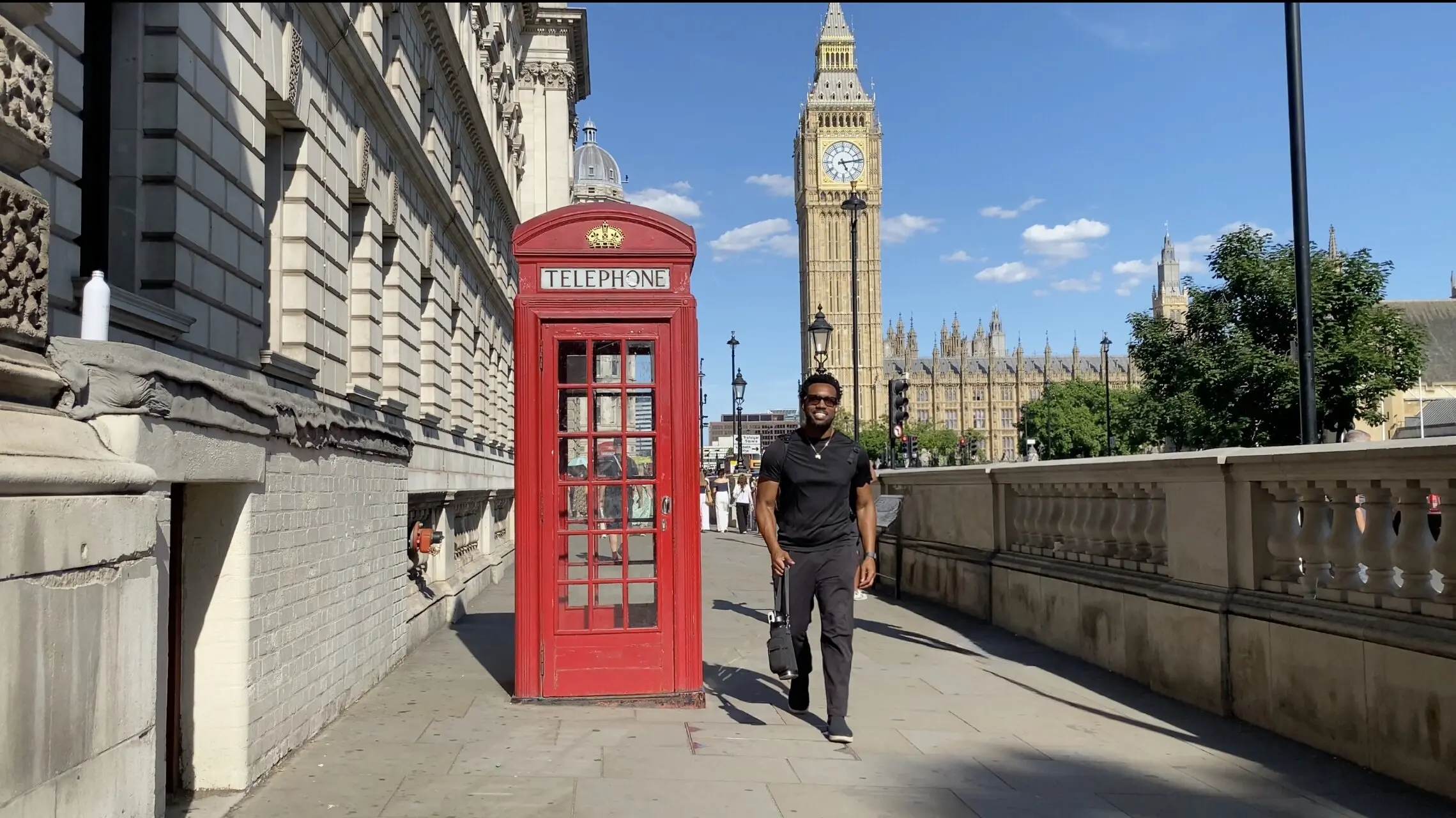 Bakari Akil walking past a red telephone box on Westminster Bridge in London, England, with Big Ben and the Houses of Parliament rising behind him under a blue sky