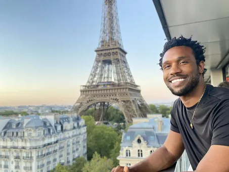 Bakari Akil smiling on a balcony at golden hour in Paris, France, the Eiffel Tower rising directly behind him above the Haussmann rooftops of the city