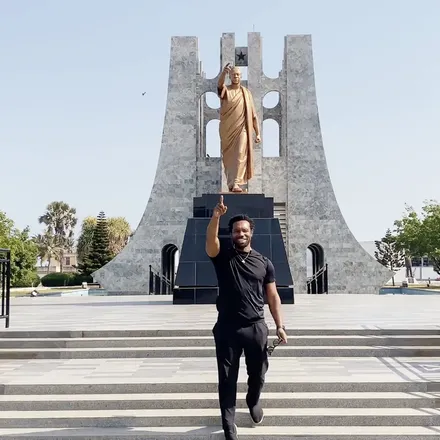 Bakari Akil pointing up at the bronze statue of Kwame Nkrumah at the Kwame Nkrumah Memorial Park in Accra, Ghana, the arched marble mausoleum rising behind the statue