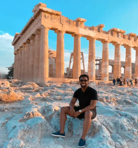 Bakari Akil in sunglasses seated on marble rocks in front of the Parthenon on the Acropolis in Athens, Greece, golden-hour light on the ancient columns