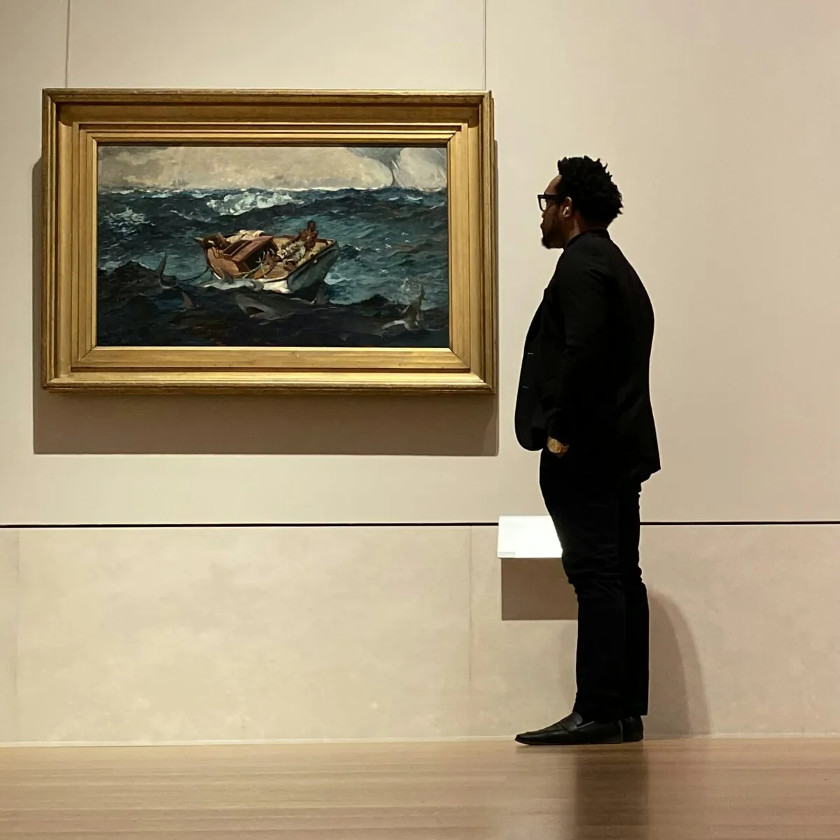 Bakari Akil, private equity investor and business acquisition lecturer, standing in profile in a museum gallery studying Winslow Homer's The Gulf Stream — a painting of a Black man alone on a broken boat surrounded by sharks — wearing all black with glasses and a gold watch