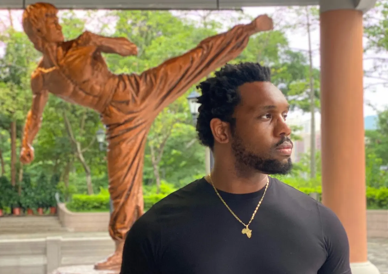 Bakari Akil standing in front of a bronze Bruce Lee high-kick statue at the Hong Kong Heritage Museum, wearing a black t-shirt and gold Africa pendant necklace, lush green trees behind him