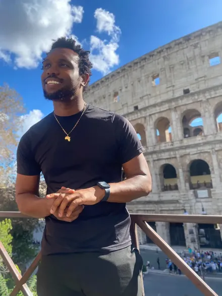 Bakari Akil in a black t-shirt and gold Africa pendant necklace smiling in front of the Colosseum in Rome, Italy, under a blue sky with white clouds