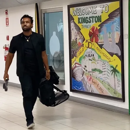 Bakari Akil in all black carrying his bags past a hand-painted Welcome to Kingston mural at the airport in Kingston, Jamaica