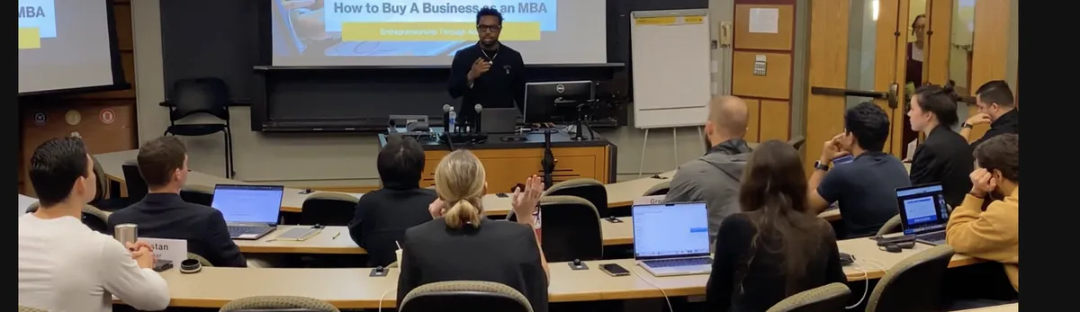 Bakari Akil lecturing on How to Buy a Business as an MBA in a packed university classroom, students with laptops raising hands, projection screen behind him