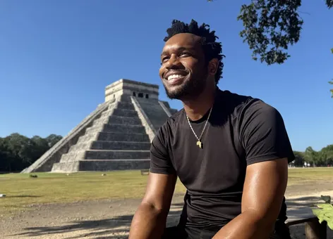 Bakari Akil smiling in the morning sun in front of El Castillo, the stepped Mayan pyramid at Chichén Itzá, Mexico, wearing a black t-shirt and gold Africa pendant necklace