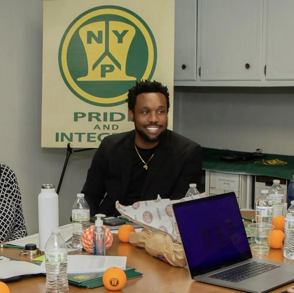 Bakari Akil smiling in the NYP Corp conference room, seated at a long table with a laptop open, wearing a black blazer and gold Africa pendant necklace, the green-and-gold NYP Pride and Integrity logo banner behind him — the $30M specialty manufacturer he acquired through Graves Hall Capital