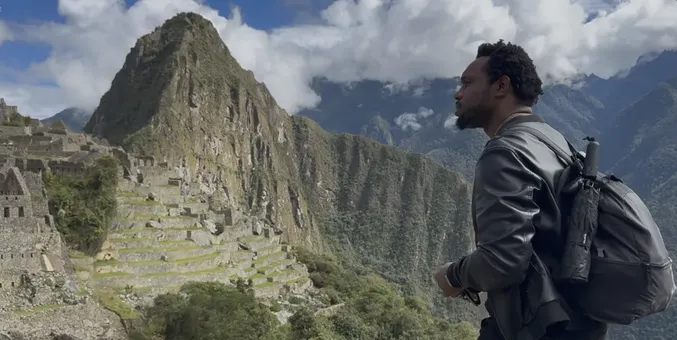 Bakari Akil in profile at Machu Picchu, Peru, wearing a black leather jacket and backpack, looking out over the ancient Inca stone terraces with Huayna Picchu rising into the clouds behind him