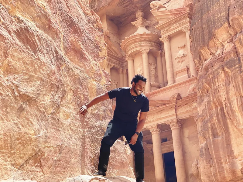 Bakari Akil posing on a rock ledge in front of Al-Khazneh, the ancient Treasury carved into rose-red sandstone cliffs at Petra, Jordan, wearing all black