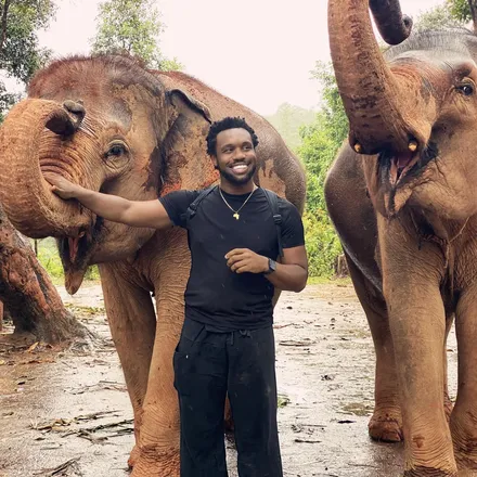 Bakari Akil smiling between two elephants at an elephant sanctuary in Thailand, wearing all black with a gold Africa pendant necklace, one elephant curling its trunk toward his outstretched hand