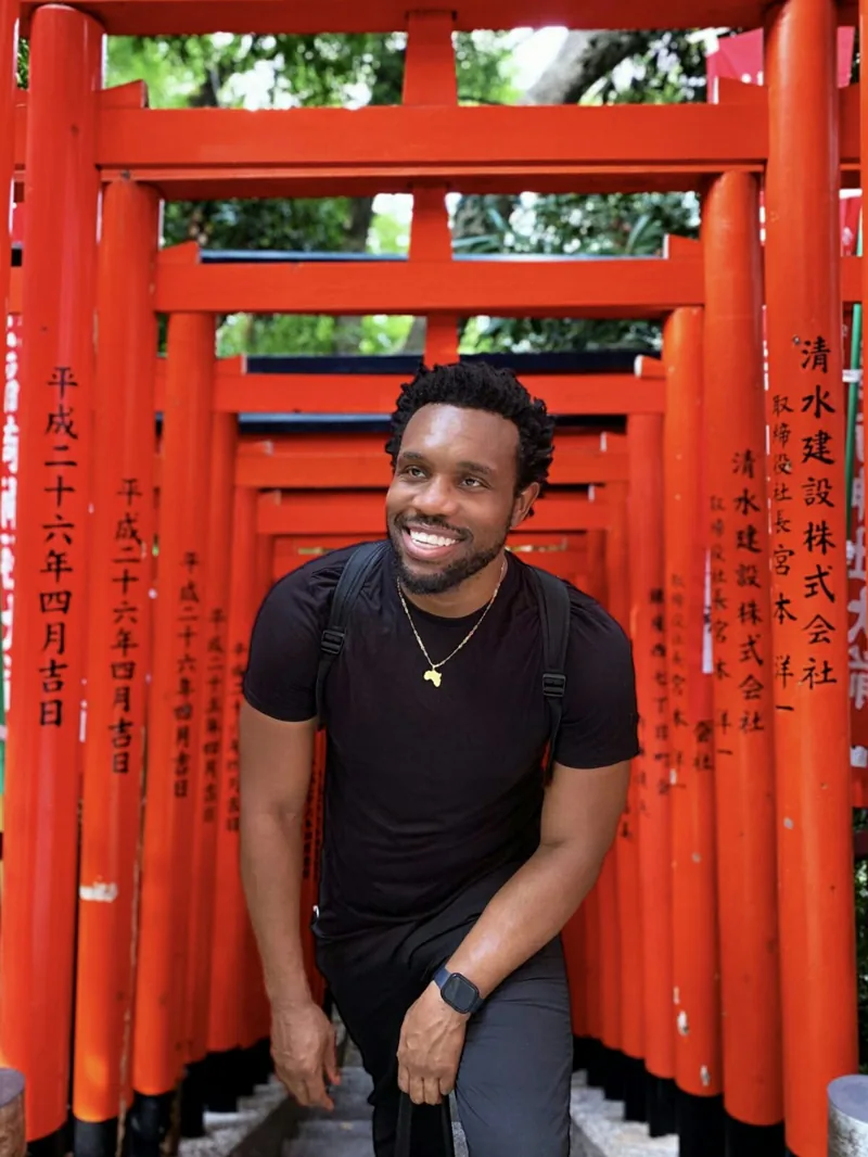 Bakari Akil laughing under a corridor of vermilion torii gates at a Shinto shrine in Tokyo, Japan, wearing a black t-shirt with a gold Africa pendant, Japanese kanji inscriptions visible on the gate pillars