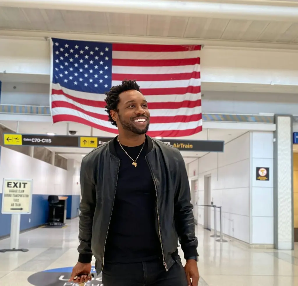 Bakari Akil smiling in a black leather bomber jacket and gold Africa pendant necklace walking through JFK Airport with a large American flag hanging overhead, Gates C70-C115 and AirTrain signs visible